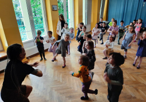 Dzieci stoją w rozsypce na sali gimnastycznej powtarzają ruchy za trenerką zumby stojąc na jednej nodze i kręcąc młynek rękami.