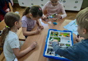 Dzieci siedzą przy stolikach i układają robociki z klocków LEGO wg. instrukcji wyświetlanej na tablecie na warsztatach z robotyki i programowania.