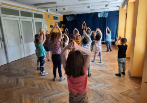 Dzieci stoją w rozsypce na sali gimnastycznej powtarzają ruchy za trenerką zumby stojąc z wyprostowanymi rękami do góry złączonymi nad głowami.