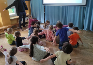Dzieci siedzą na podłodze, oglądają prezentację nt. kina niemego i machają rękami wystawionymi do boku za prowadzącym warsztaty.