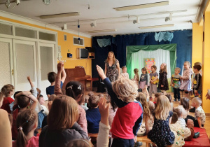 Prowadząca koncert recytuje wiersze, a dzieci stoją wymachując rękami na boki i wykonują polecenia, jakie im zleca.