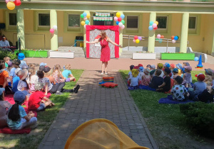 Dzieci siedzą na kocach rozłożonych na trawie w ogrodzie przedszkolnym, prowadząca koncert śpiewa im piosenki z repertuaru stojąc na środku z rozłożonymi rękami do boku, a Pan na keyboardzie jej akompaniuje.