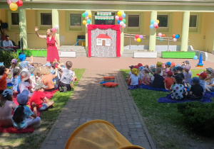 Dzieci siedzą na kocach rozłożonych na trawie w ogrodzie przedszkolnym, prowadząca koncert śpiewa im piosenki z repertuaru machając do dzieci, a Pan na keyboardzie jej akompaniuje.