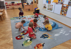 Na dywanie rozsypane leżą kolorowe listeczki z papieru, dzieci je zbierają.