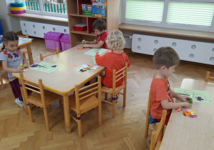 Trzech chłopców i dziewczynka siedzi przy stoliku, naklejają na sylwetę drzewa zebrane wcześniej listki.