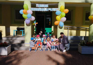 Cała grupa siedzi na schodach przed budynkiem przedszkola wraz z wychowawcą, nad nimi wisi kolorowy napis ,,Dzień Przedszkolaka" oraz baloniki.