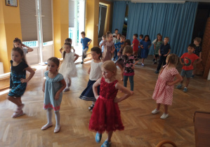 Dzieci stoją w rozsypce na sali gimnastycznej, trzymają ręce na biodrach i uczą się układu tanecznego do piosenki.