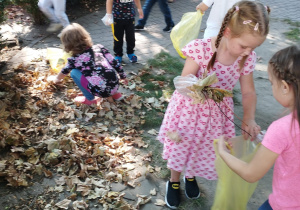 Jedna z dziewczynek trzyma worek przed sobą, natomiast druga wkłada do niego zebrane liście.