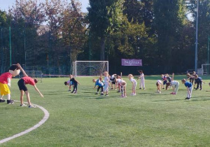 Przedszkolaki wraz z Varsikiem oraz trenerami wykonują skręty tułowia w ramach rozgrzewki.