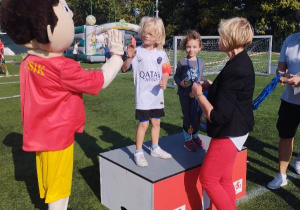 Na podium stoi chłopiec z dziewczynką, którzy wygrali bieg. Chłopiec przybija piatkę z Varsikiem, a dziewczynka pozuje z otrzymanym medalem.