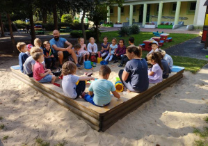 Cała grupa wraz z prowadzącymi warsztaty siedzi na brzegach piaskownicy, dzieci słuchają wprowadzenia do tematu zajęć.