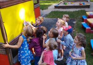 Dzieci stoją przed domkiem na placu zabaw i patrzą na maskotkę, którą trzyma w ręku jeden z prowadzących warsztaty.