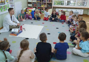Prowadzący zajęcia siedzi na dywanie, przed sobą ma rozłożoną białą ceratę, na której znajdują się rekwizyty związane z tematyką zajęć. Dzieci siedzą w półkolu na dywanie.