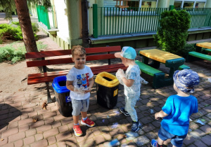 Trzej chłopcy stoją przy pojemnikach do segregacji odpadów, jeden z nich jest na papier, a drugi na plastik.