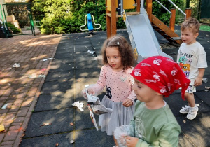 Dziewczynka oraz dwaj chłopcy idą w kierunku kosza na papier, aby wyrzucić do niego zebrane w ogrodzie papierowe śmieci.