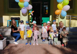 Przed wejściem do budynku stoją dzieci wraz z Paniami, a nad nimi wiszą kolorowe balony oraz napis: ,,Dzień Przedszkolaka".