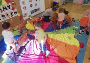 Na dywanie rozłożona jest kolorowa chusta, na której rozsypane są kasztany i żołędzie. Dzieci zbierają je i segregują do osobnych koszyków.