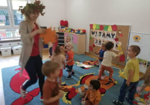 Dzieci wraz z Panią tańczą na dywanie w rytm jesiennej piosenki, w ręku trzymają kolorowe listki.