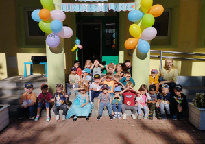 Cała grupa siedzi na schodach przed budynkiem przedszkola wraz z wychowawcą, nad nimi wisi kolorowy napis ,,Dzień Przedszkolaka" oraz baloniki.