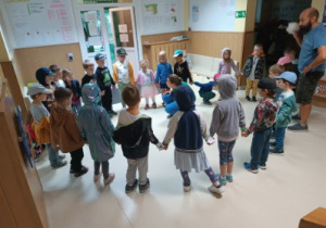 Dzieci stoją w holu budynku, tworzą koło zamknięte trzymając się za ręce. W środku koła kuca Pani prowadząca warsztaty.