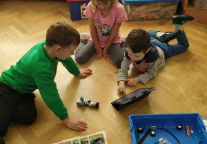 Dwóch chłopców i dziewczynka siedzą na parkiecie i patrzą na instrukcję w tablecie, przed nimi znajduje się budowany z klocków robot.