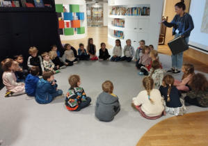 Dzieci siedzą na dywanie w Muzeum POLIN i słuchają Pani, która wprowadza ich w tematykę warsztatów.