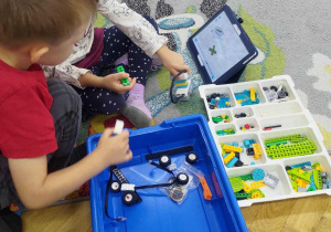 Dziewczynka i chłopiec składają elementy robota według instrukcji na tablecie.
