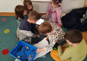 Trzy dziewczynki i trzech chłopców próbują zbudować robota. Jeden z chłopców szuka w zestawie odpowiedniej części.