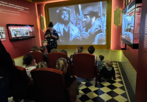 Dzieci siedzą w fotelach i oglądają film w niemym kinie.