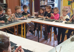 Dzieci siedzą za stołami ustawionymi w podkowę i lepią figurki z gliny.