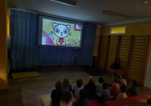 Na ekranie w sali gimnastycznej wyświetlany film o Kici Koci oglądają dzieci z oddziału I.