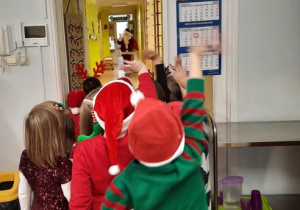 Dzieci z oddziału III stoją w drzwiach sali i machają do Mikołaja znajdującego się w holu przedszkola.