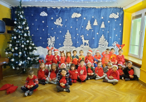 Dzieci z grupy III siedzą przy choince na sali gimnastycznej, w dłoniach trzymają otrzymane od Mikołaja upominki.