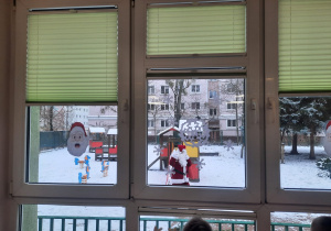 Dzieci wyglądają przez okno, pod którym spaceruje Mikołaj dzwoniący dzwonkiem i machający do przedszkolaków.