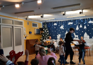 Dwie pary dzieci i artysta z chłopcem tańczą na środku sali do muzyki granej na żywo przez muzyków.