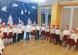 Starszaki stoją w półkolu, trzymając się za ręce śpiewają świąteczną piosenkę.
