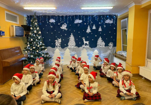 Przedszkolaki siedzą na podłodze w czterech rzędach, a na głowach mają świąteczne czapeczki.