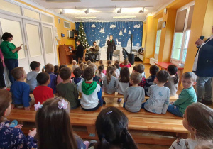 Dzieci siedzą na ławeczkach, a na środku artyści prezentują za pomocą muzyki i śpiewu program artystyczny.