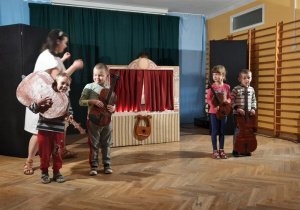 Na środku stoją trzej chłopcy oraz dziewczynka w ręku trzymając instrumenty smyczkowe wykorzystywane w operowej orkiestrze.