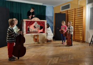 Czworo dzieci z instrumentami stoi zwrócone w stronę sceny, na której występuje marionetkowy dyrygent.