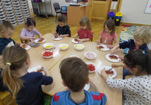 Dzieci siedzą przy złączonych stolikach i smarują sos pomidorowy na mini pizzach.