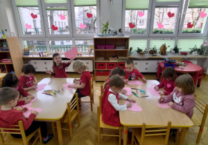 Przedszkolaki siedzą przy stolikach i ozdabiają kredkami serduszka.