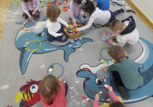 Dzieci siedzą na dywanie, przed nimi leżą krążki gimnastyczne, wokół których układają figury geometryczne.