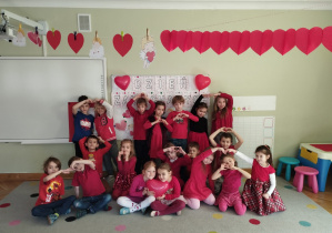 Cała grupa ubrana w czerwone ubrania pozuje do wspólnego zdjęcia z okazji Dnia zakochanych, za pomocą rąk tworzą małe i duże serduszka.