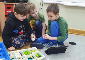 Jeden z chłopców trzyma w ręku klocki, drugi ma w dłoni kolejną część do zbudowania robota, a trzeci sprawdza zadanie w tablecie.
