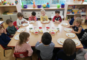 Cała grupa siedzi przy złączonych stolikach, przed każdym dzieckiem leży talerzyk z chlebkiem, który za moment będą ozdabiać wybranymi przez siebie produktami.