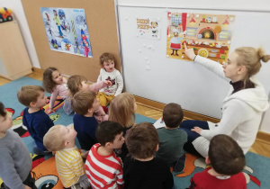 Dzieci siedzą przed tablicą, na której zawieszony jest plakat ukazujący wnętrze pizzerii i omawiają je z nauczycielką.
