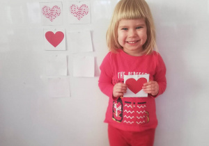 Dziewczynka stoi przy tablicy, w ręku trzyma ilustrację serduszka identyczną z zawieszoną na tablicy.