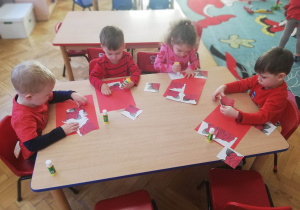 Trzech chłopców i dziewczynka siedzą przy jednym stoliku i naklejają na kartki układankę z 4 elementów.