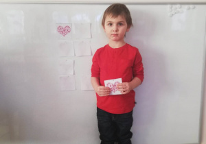 Chłopiec stoi przy tablicy, w ręku trzyma ilustrację serduszka identyczną z zawieszoną na tablicy.